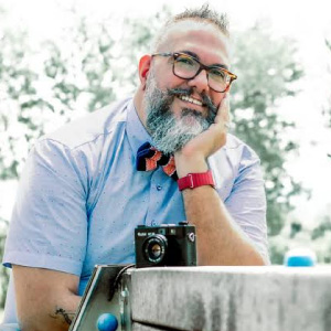 Foto in primo piano di Mauro Santoro , uomo sorridente con occhiali, baffi e barba