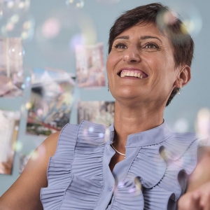 Primo piano di donna sorridente, capelli corti e blusa lilla, contornata da bolle di sapone.